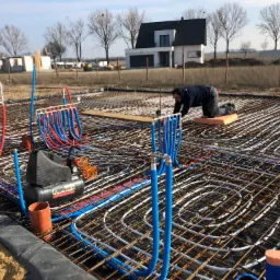 Instalacja rur ogrzewania podłogowego w siatce zbrojeniowej, z widocznym kompresorem powietrza i pracownikiem na kolanach, w tle budowa domu jednorodzinnego.