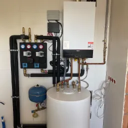 Nowoczesny piec gazowy Viessmann zamontowany na białej ścianie z płytkami, z widoczną instalacją hydrauliczną i czarnymi rurami izolowanymi, oraz białym zasobnikiem ciepłej wody, częściowo zakrytym...