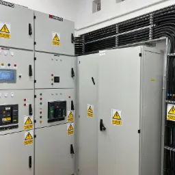 Szare szafy elektryczne z ostrzeżeniami 'Nie dotykać! Urządzenie elektryczne!' w pomieszczeniu z rurami instalacyjnymi przy suficie i kratkami wentylacyjnymi.