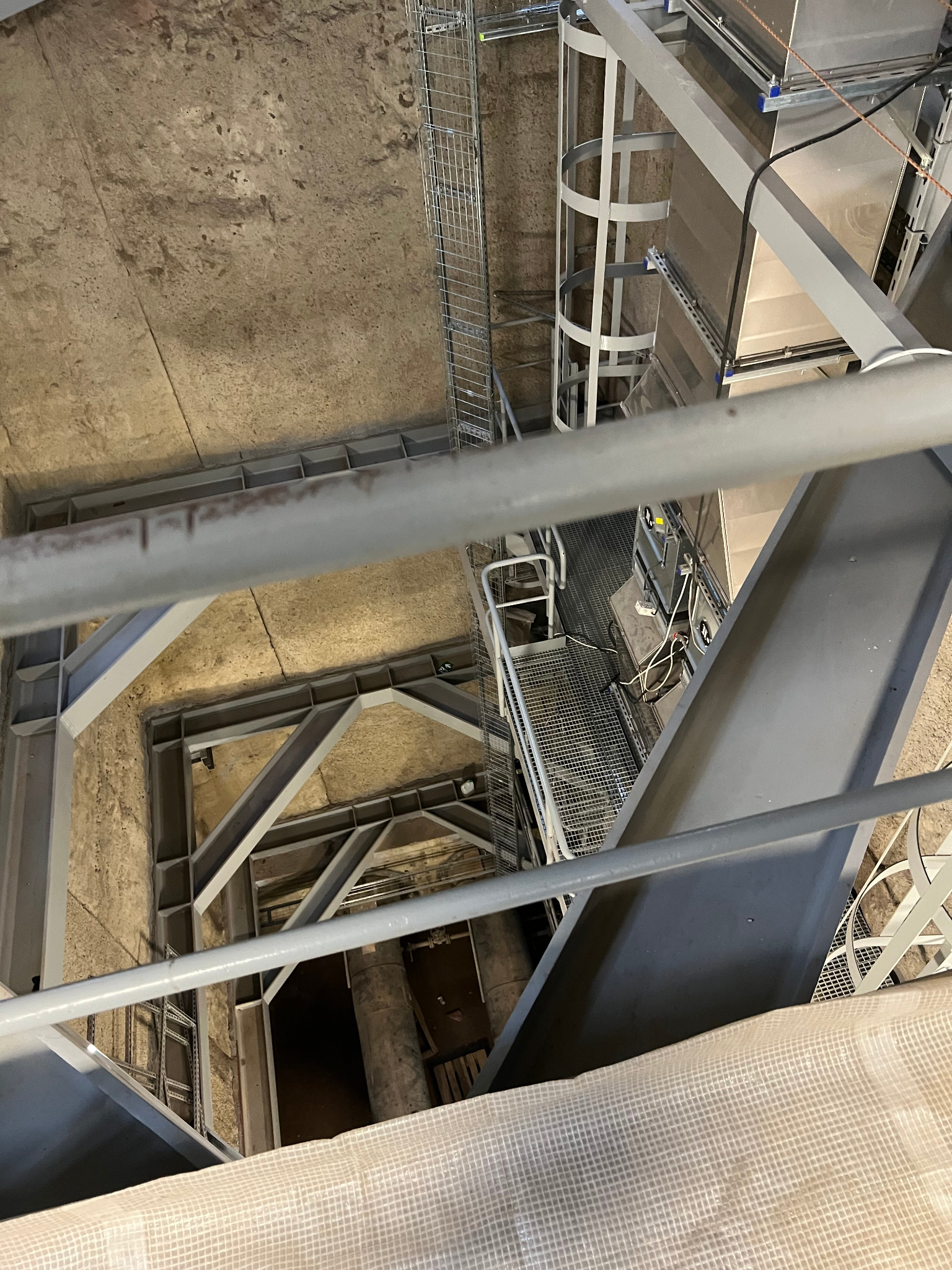 Wnętrze techniczne z metalowymi konstrukcjami, drabinkami i instalacjami, widok z góry na betonowe ściany i podłogi, zabezpieczone częściowo białą plandeką.