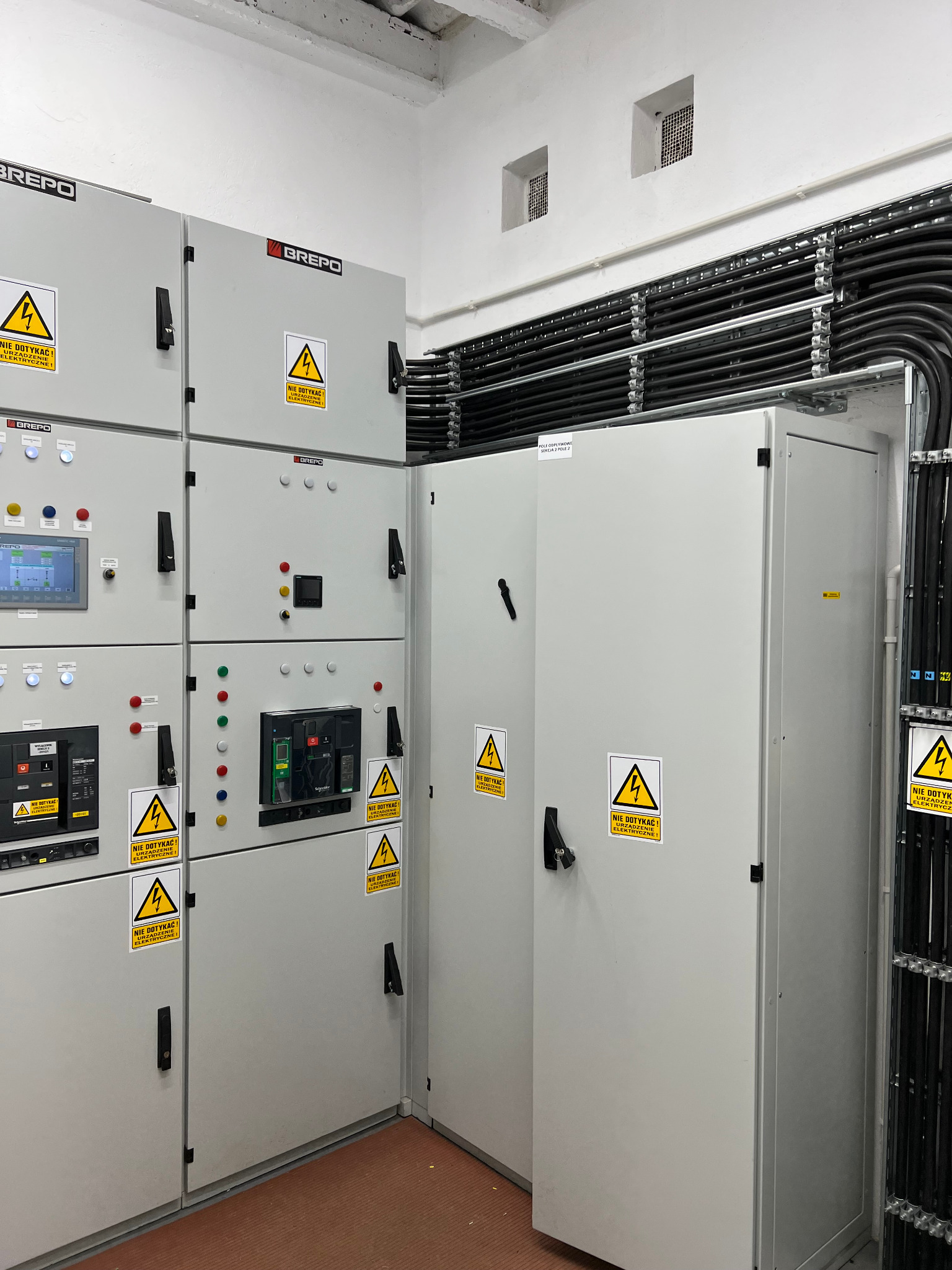 Szare szafy elektryczne z ostrzeżeniami 'Nie dotykać! Urządzenie elektryczne!' w pomieszczeniu z rurami instalacyjnymi przy suficie i kratkami wentylacyjnymi.