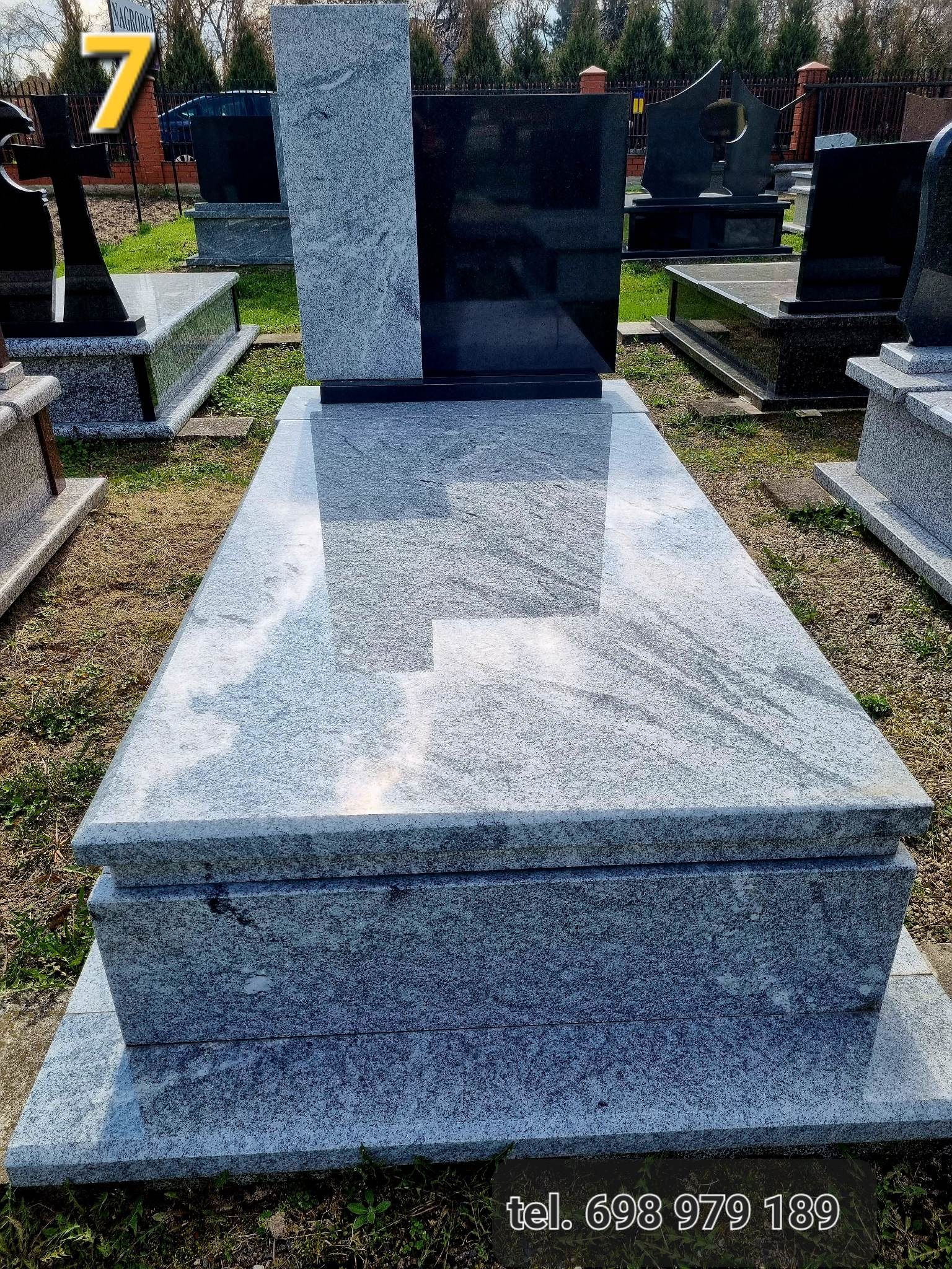 Szary, polerowany nagrobek z granitu z czarnym elementem pionowym w tle, widoczny na cmentarzu w słoneczny dzień.