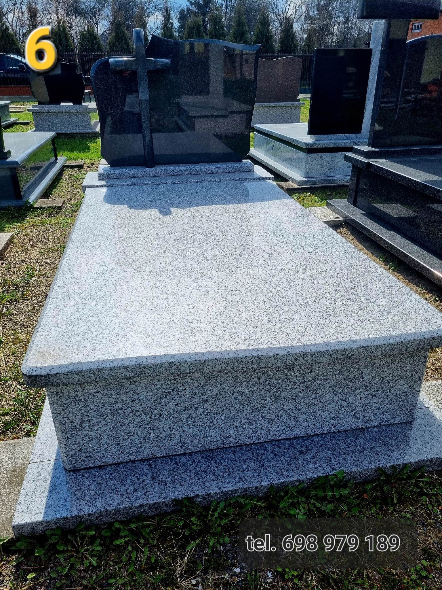 Szary nagrobek z ciemnym krzyżem na cmentarzu, widoczne inne nagrobki w tle.