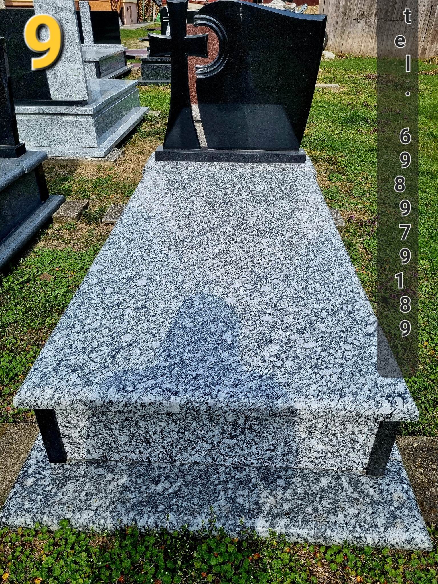 Granitowy nagrobek z czarnym krzyżem w tle, otoczony trawą i innymi nagrobkami.