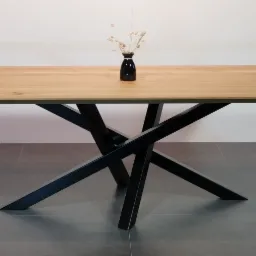 Stół z blatem z jasnego drewna i geometryczną podstawą z czarnych, skrzyżowanych metalowych profili, na blacie czarny wazon z suszonymi kwiatami, całość na tle szarej podłogi i białej ściany.