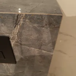 Fragment zabudowy geberit w łazience wykończonej płytami imitującymi szary marmur z wyraźnym użyleniem, widoczny przycisk spłukujący w kolorze czarnym.