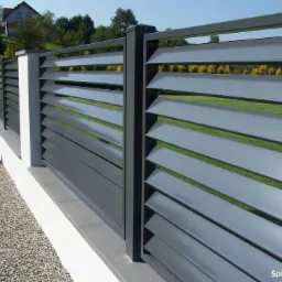 Nowoczesne ogrodzenie posesji z poziomych, szarych paneli metalowych, osadzonych na białym, niskim murku z kamienną podsypką i słupkami.