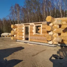 Surowa konstrukcja ścian z bali z widocznymi otworami okiennymi i drzwiowymi, w tle las i fragment żurawia budowlanego.