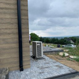 Krystian Łukasik - Ułożona szara kostka brukowa przy elewacji budynku z zamontowanym odpływem rynnowym i jednostką zewnętrzną klimatyzacji, widok na okoliczne wzgórza i zachmurzone niebo.