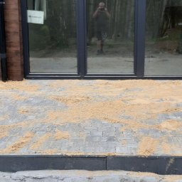 Krystian Łukasik - Ułożona szara kostka brukowa, częściowo zasypana piaskiem, przed budynkiem z drewnianą elewacją i dużymi oknami z ciemnymi ramami; w odbiciu okien widać osobę robiącą zdjęcie.