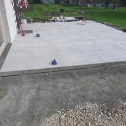 Krystian Łukasik - Jasnoszare płytki tarasowe ułożone na zewnątrz budynku, widoczne narzędzia do przenoszenia płytek i przygotowane podłoże z kamienia i betonu.