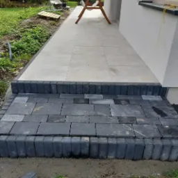 Ułożone stopnie z ciemnej kostki brukowej prowadzące do jasnego, wyłożonego płytkami tarasu przy domu jednorodzinnym. Widoczny fragment ogrodu z trawą i huśtawką w tle.