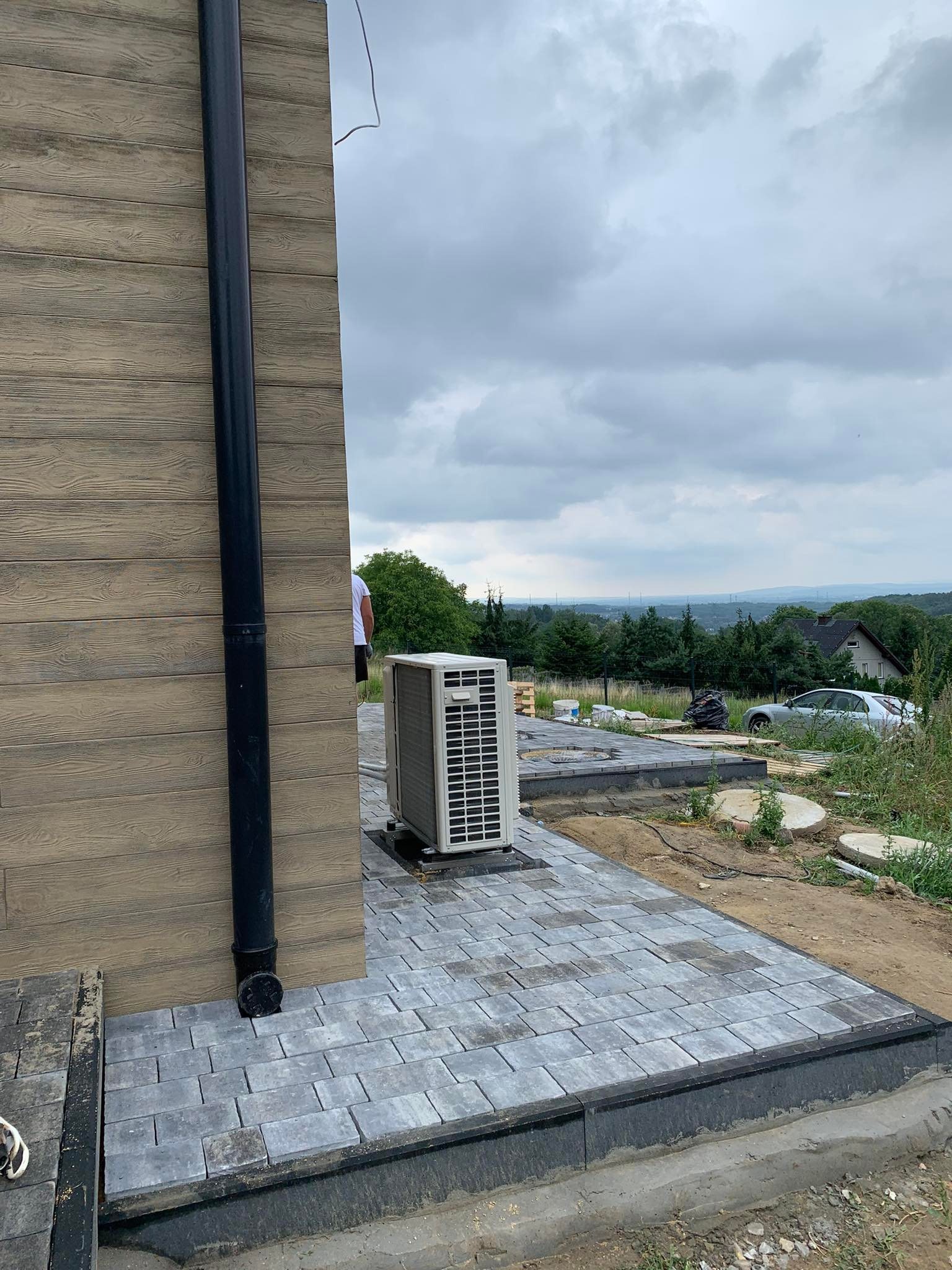 Ułożona szara kostka brukowa przy elewacji budynku z zamontowanym odpływem rynnowym i jednostką zewnętrzną klimatyzacji, widok na okoliczne wzgórza i zachmurzone niebo.