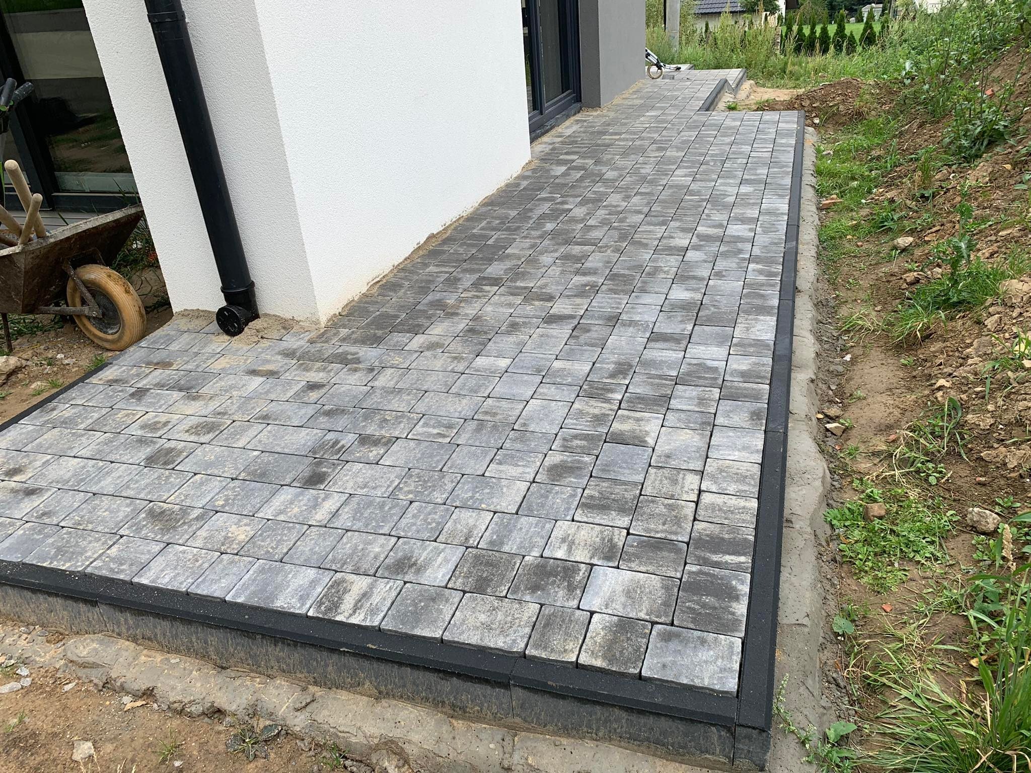 Szara kostka brukowa ułożona przy białej ścianie budynku, widoczny czarny obrzeże i taczka z narzędziami w tle.