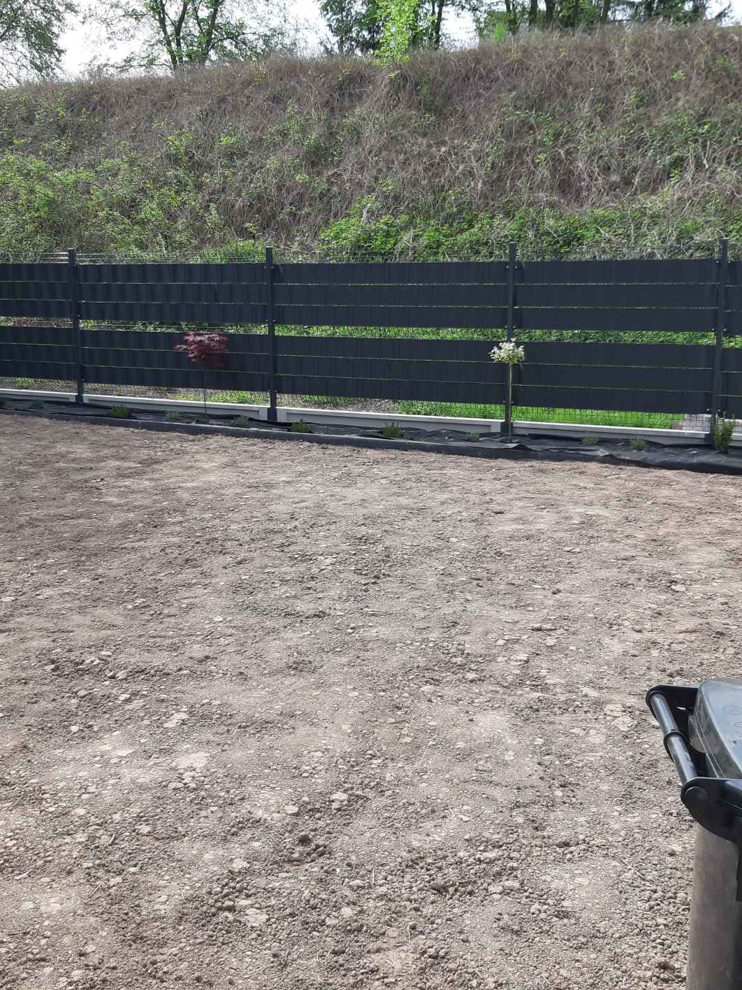 Nowoczesne, poziome, grafitowe panele ogrodzeniowe zamontowane na metalowych słupkach, widoczne małe drzewka wzdłuż ogrodzenia, teren przygotowany pod przyszłe prace brukarskie.