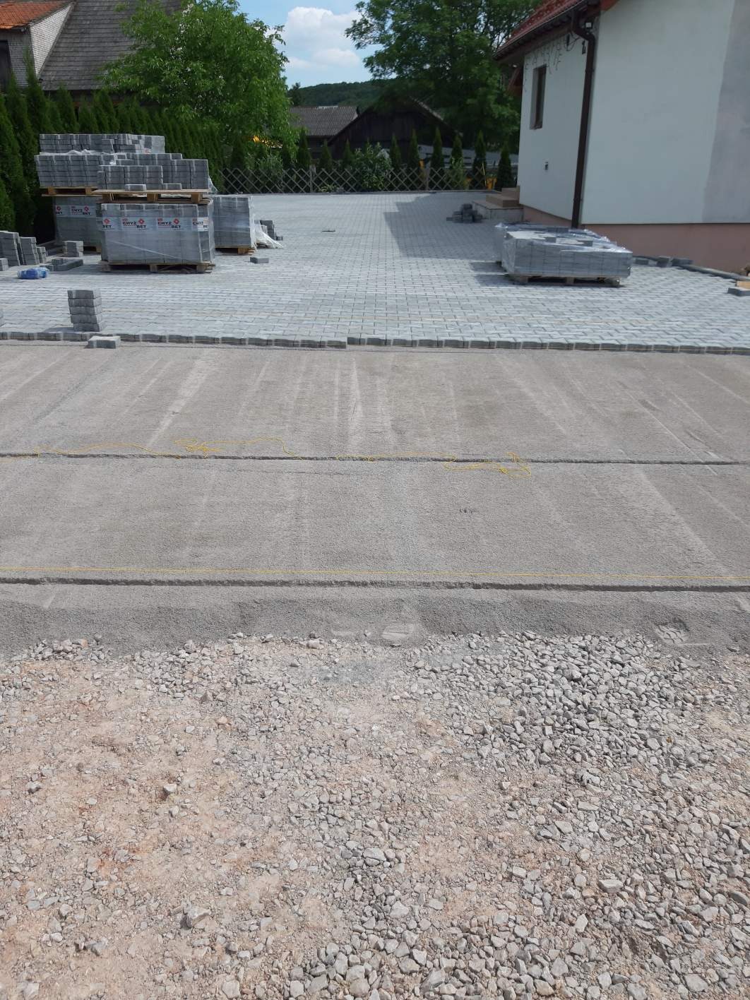 Przygotowanie podłoża pod nawierzchnię z kostki brukowej: warstwa podsypki piaskowo-cementowej, obrzeża ułożone, palety z kostką w tle, widok z góry.