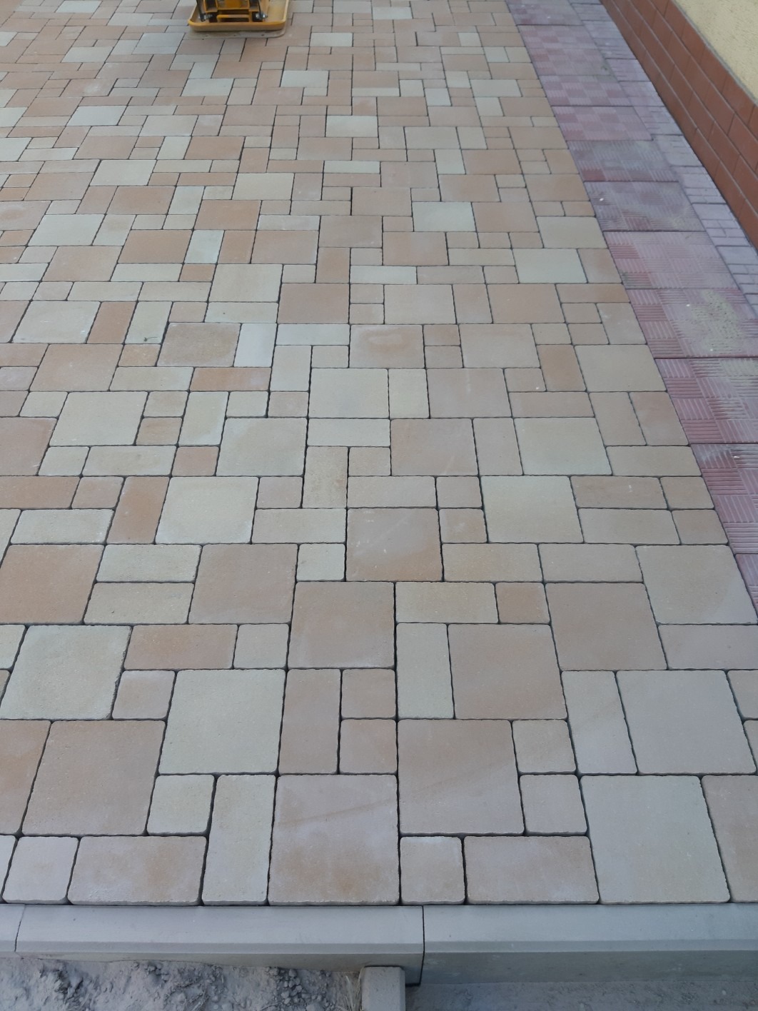 Nowo ułożona, beżowo-brązowa kostka brukowa o nieregularnym wzorze, częściowo ubijana zagęszczarką w górnym kadrze, z widocznym obrzeżem i fragmentem ściany z cegły po prawej stronie.