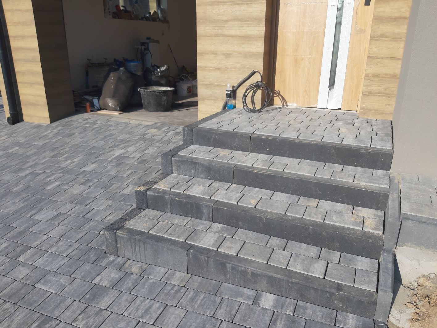 Schody wejściowe wyłożone szarą kostką brukową, prowadzące do drewnianych drzwi z przeszkleniem, widok na garaż z workami i wiadrem w tle.