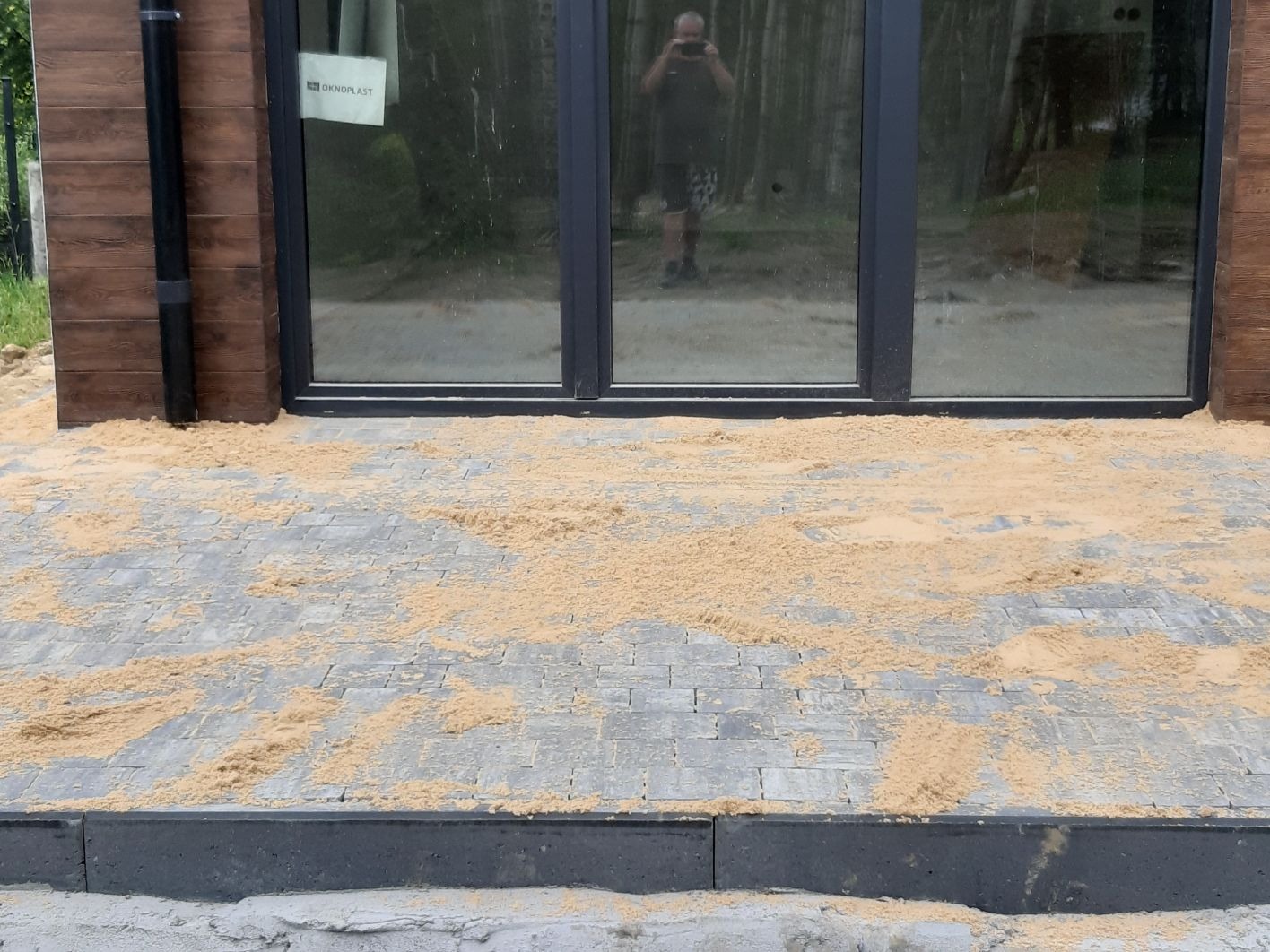 Ułożona szara kostka brukowa, częściowo zasypana piaskiem, przed budynkiem z drewnianą elewacją i dużymi oknami z ciemnymi ramami; w odbiciu okien widać osobę robiącą zdjęcie.