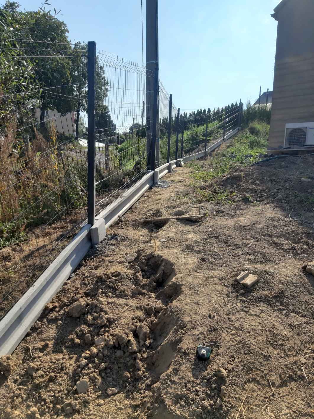 Montaż ogrodzenia panelowego na betonowej podmurówce, widoczne słupki i siatka, teren nierówny, w tle budynek i zieleń.