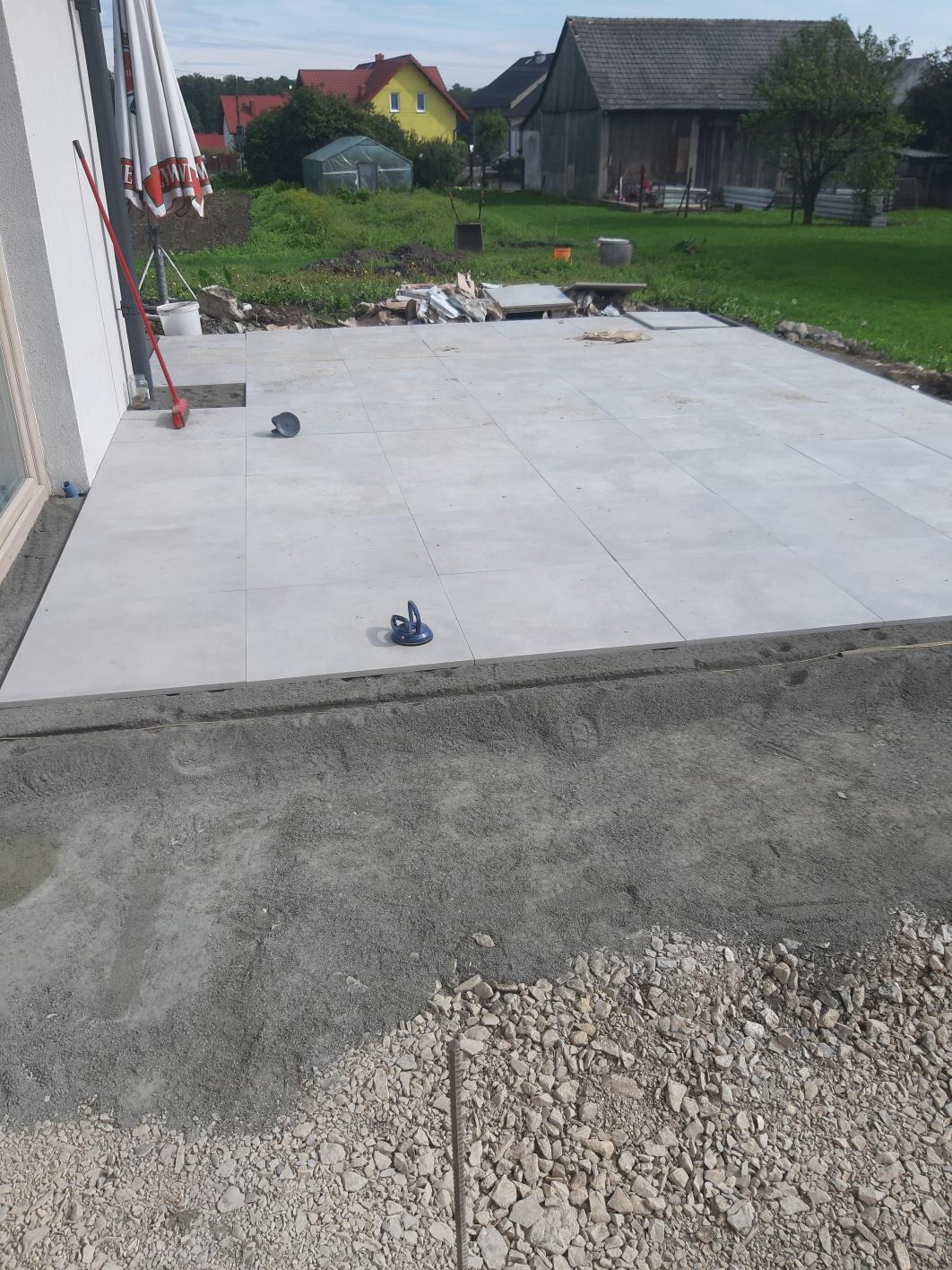 Jasnoszare płytki tarasowe ułożone na zewnątrz budynku, widoczne narzędzia do przenoszenia płytek i przygotowane podłoże z kamienia i betonu.