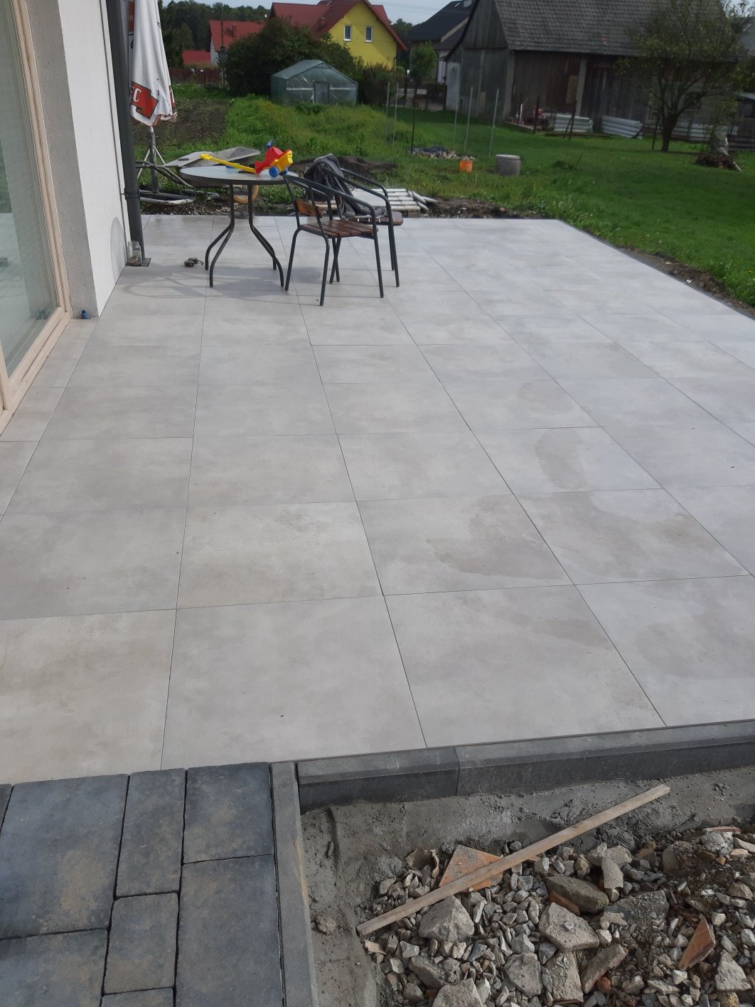Jasnoszary taras z płytek, widoczne przygotowania do ułożenia obrzeży z kostki, w tle dom z ogrodem.