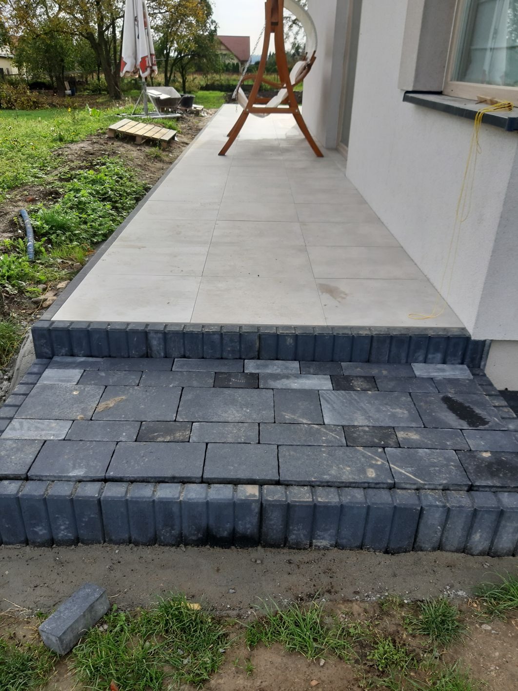 Ułożone stopnie z ciemnej kostki brukowej prowadzące do jasnego, wyłożonego płytkami tarasu przy domu jednorodzinnym. Widoczny fragment ogrodu z trawą i huśtawką w tle.