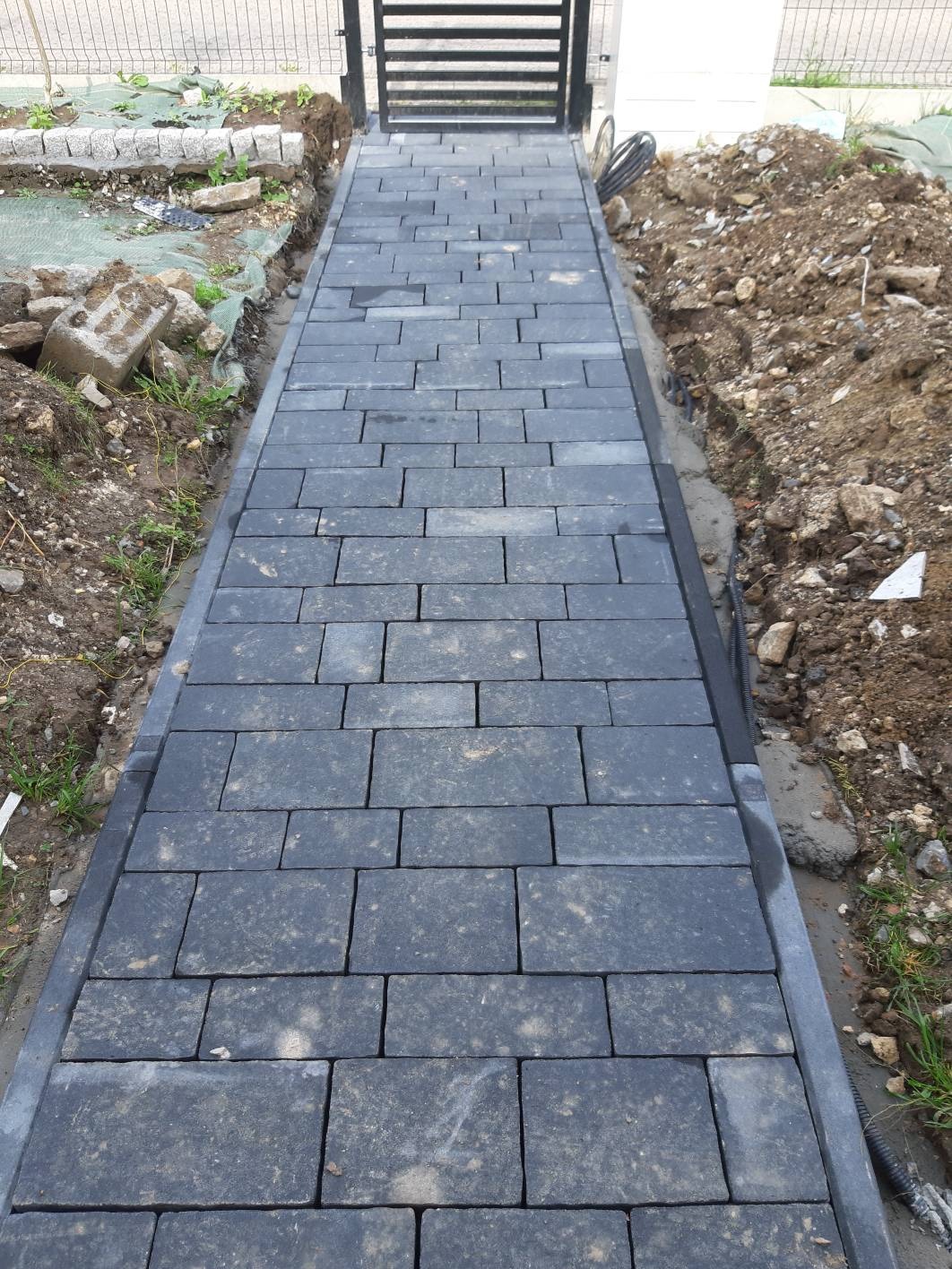 Ścieżka z ciemnoszarej kostki brukowej prowadząca do czarnej, metalowej bramki, widoczne niedokończone prace ziemne po bokach.