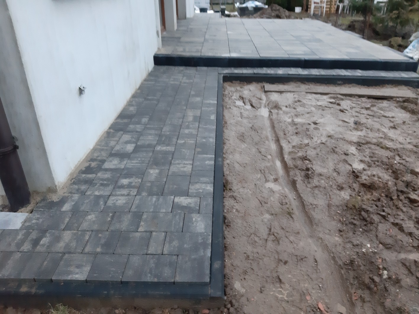 Ułożona, grafitowa kostka brukowa tworząca ścieżkę i taras przy budynku, z widocznym fragmentem terenu przygotowanego pod dalsze prace.