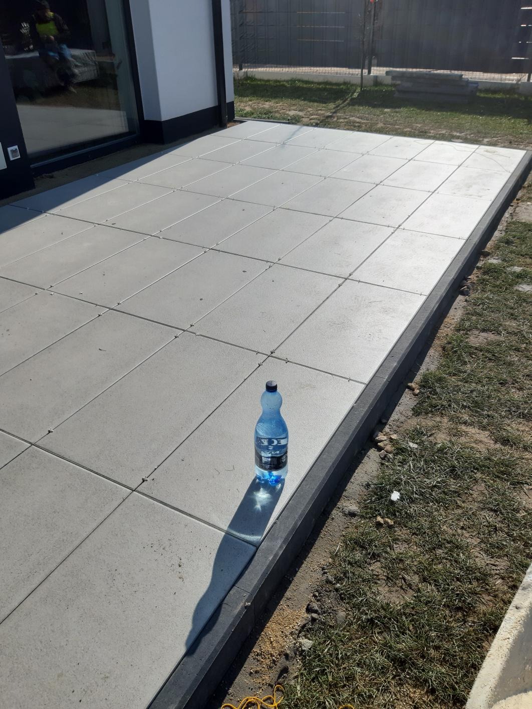 Nowo ułożony taras z szarych płyt betonowych obramowany ciemnoszarą obwódką, na jednej z płyt stoi butelka wody mineralnej.