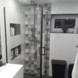 Nowoczesna łazienka z kabiną prysznicową typu walk-in z czarnymi profilami, wzorzystymi płytkami w strefie prysznica, białą umywalką i czarnym koszem na pranie.