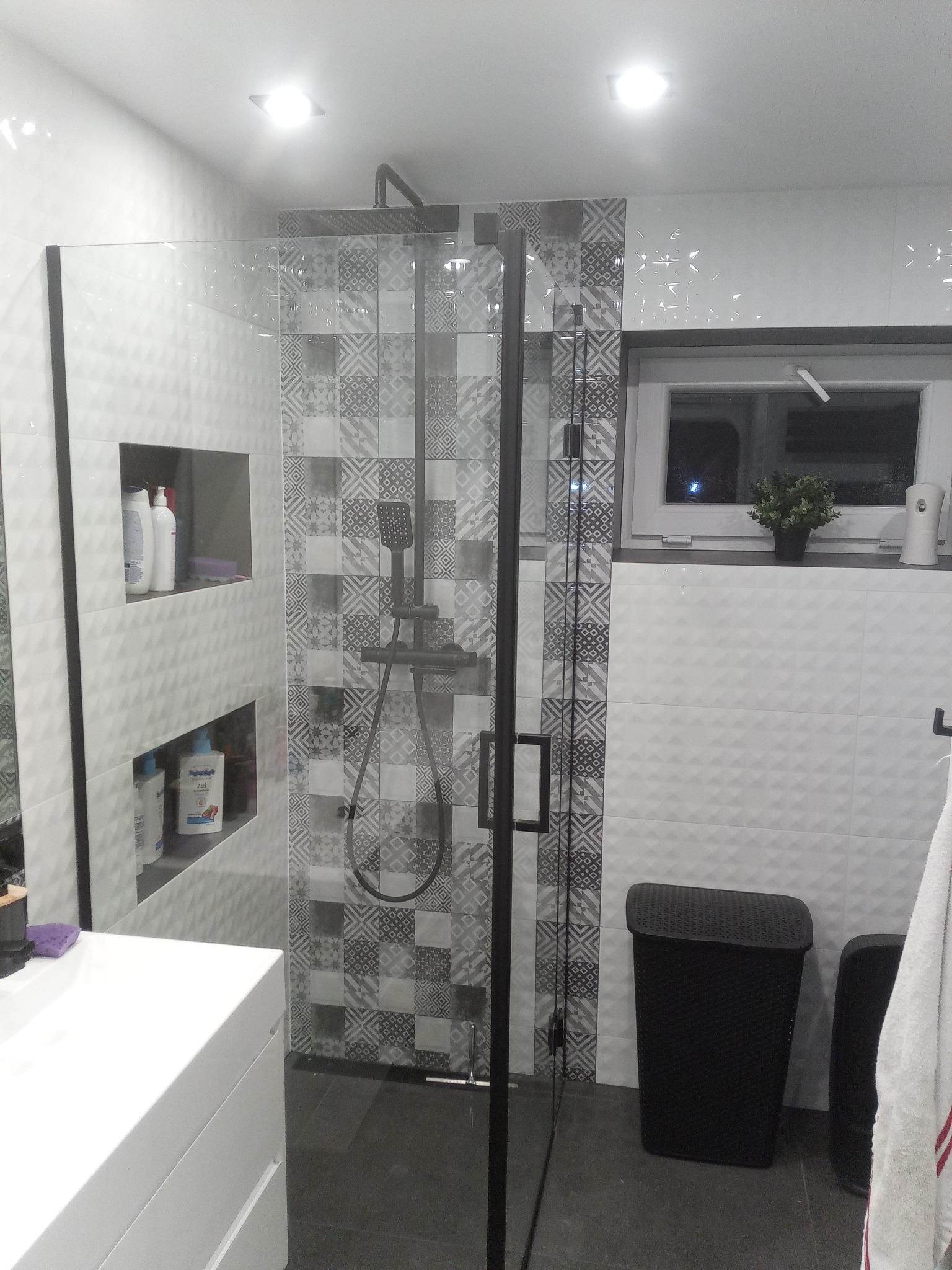 Nowoczesna łazienka z kabiną prysznicową typu walk-in z czarnymi profilami, wzorzystymi płytkami w strefie prysznica, białą umywalką i czarnym koszem na pranie.