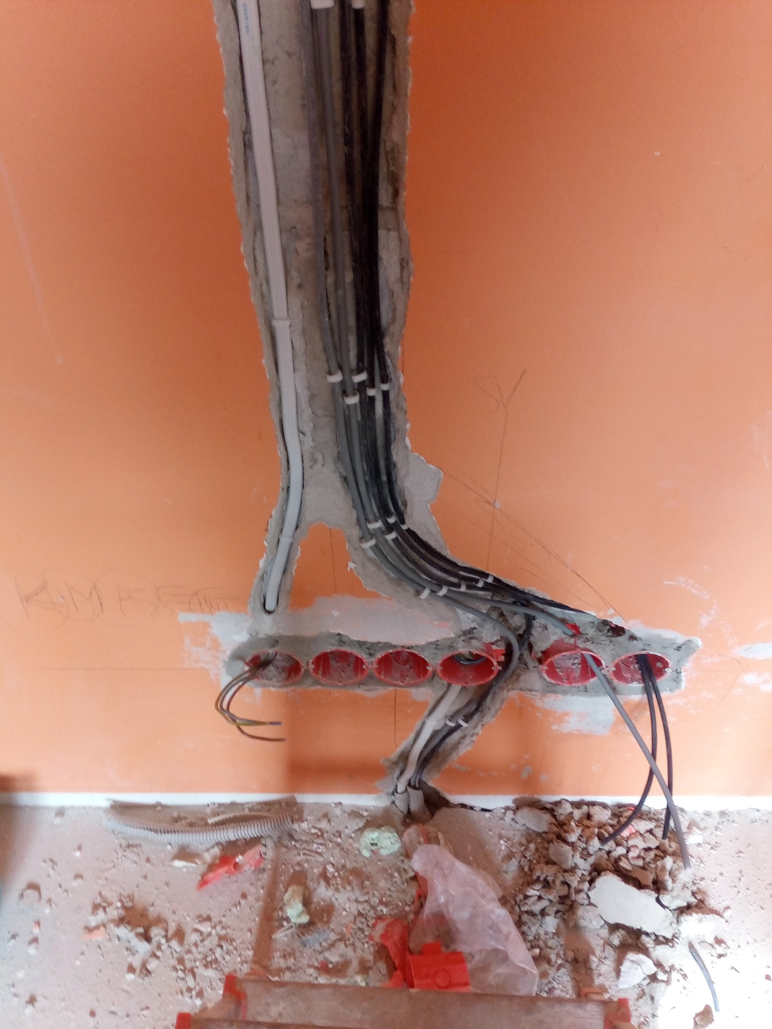 Instalacja elektryczna w ścianie z pomarańczową farbą, widoczne przewody w peszlu i puszki elektryczne, gruz na podłodze.