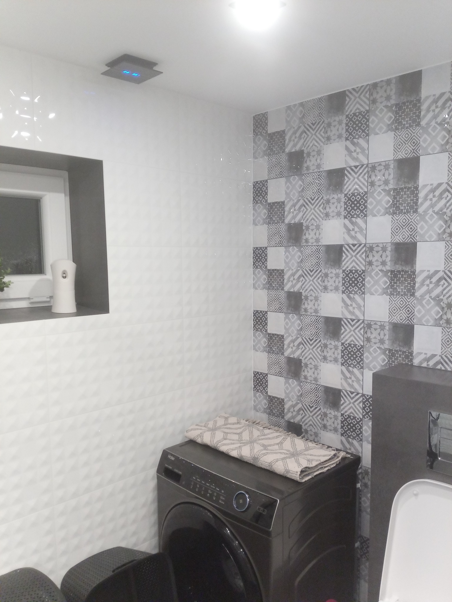 Nowoczesna łazienka z pralką automatyczną, patchworkowymi płytkami w odcieniach szarości i białymi trójwymiarowymi płytkami ściennymi, uzupełniona o okno z dyfuzorem zapachu i sufitowy panel LED.