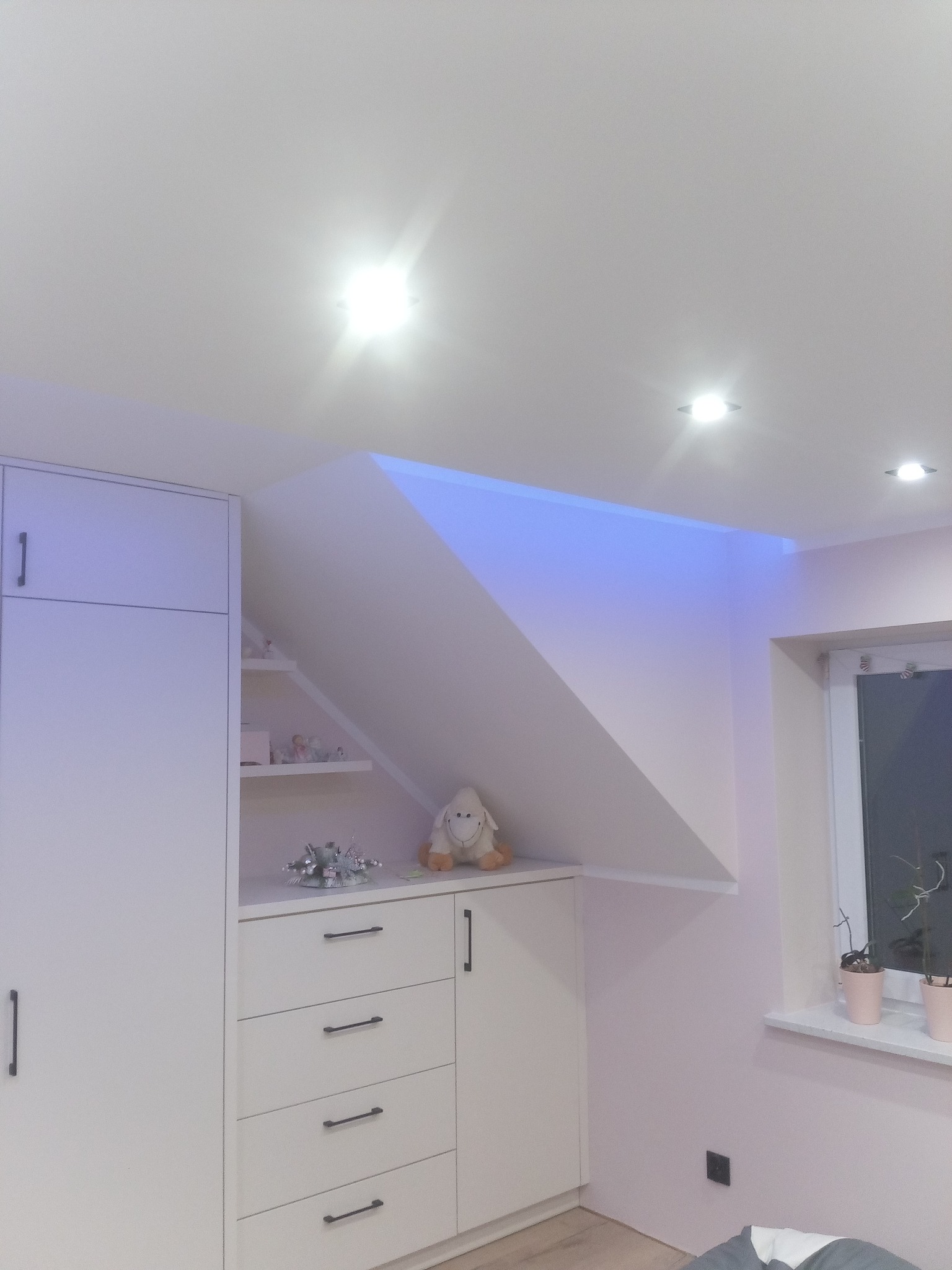 Biała zabudowa meblowa na poddaszu ze skosami, oświetlenie LED w kolorze niebieskim, zabawka pluszowa na półce, okno z roślinami w doniczkach.