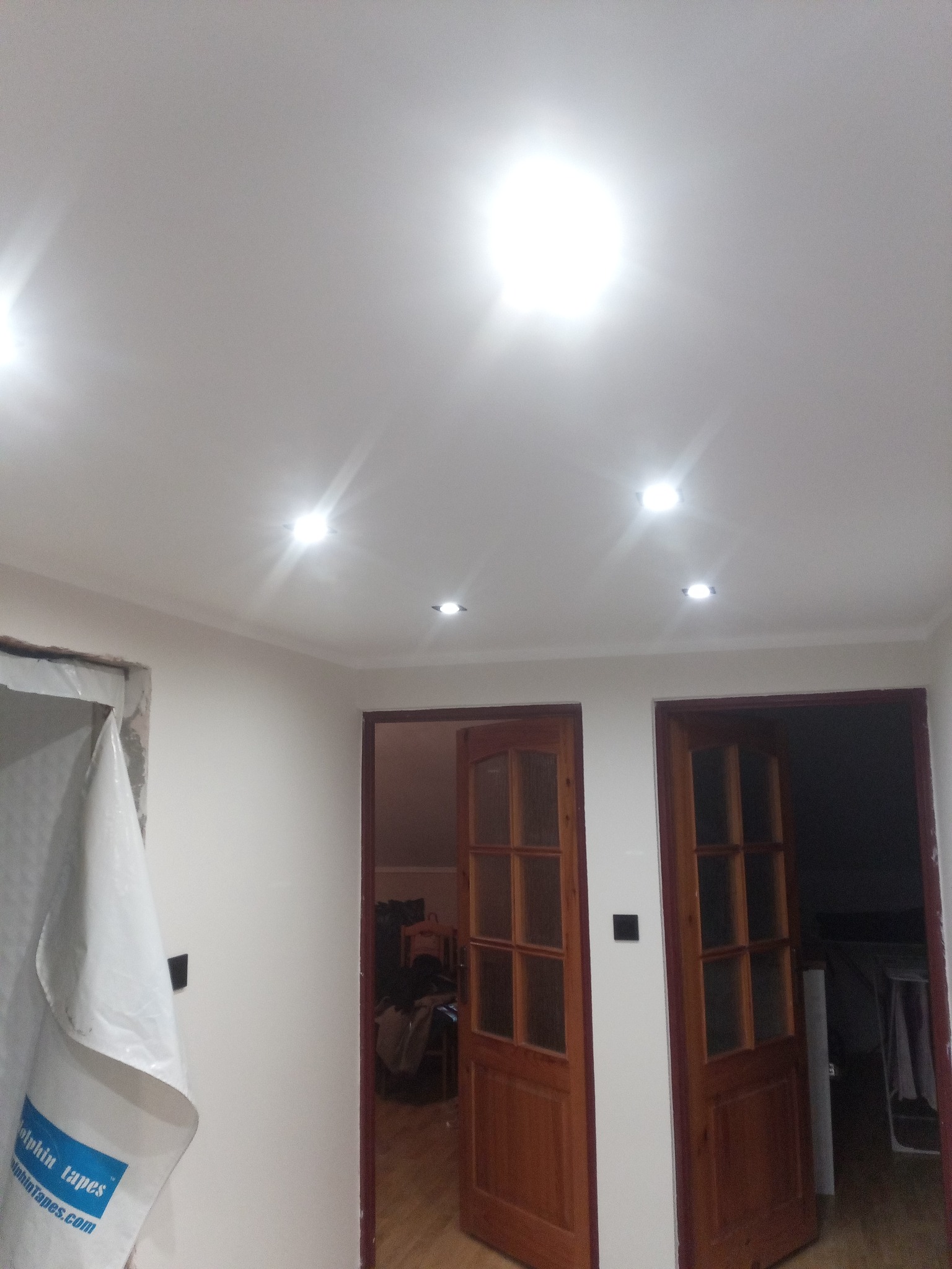 Wykończone wnętrze z pomalowanymi ścianami na biało, dwoje drewnianych drzwi z szybkami, w suficie zamontowane halogenowe lampki, zabezpieczona folią malarską framuga drzwi.