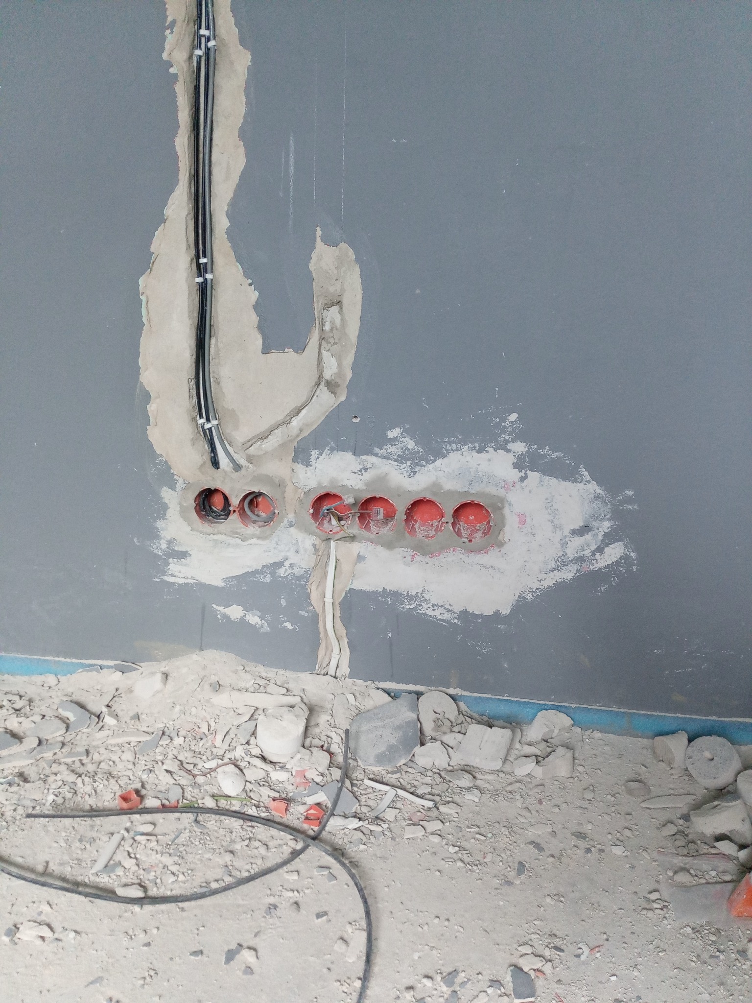 Szara ściana w trakcie remontu z wywierconymi otworami pod puszki elektryczne, wystającymi kablami i gruzem na podłodze.