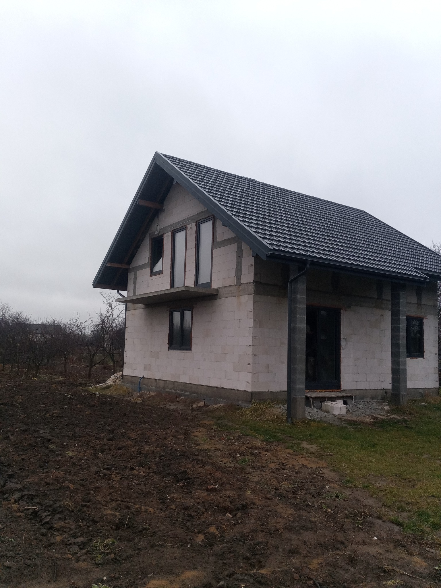 Niedokończony dom z białego pustaka z ciemną dachówką i czarnymi oknami w pochmurny dzień, widoczne fundamenty i częściowo zagospodarowany teren.