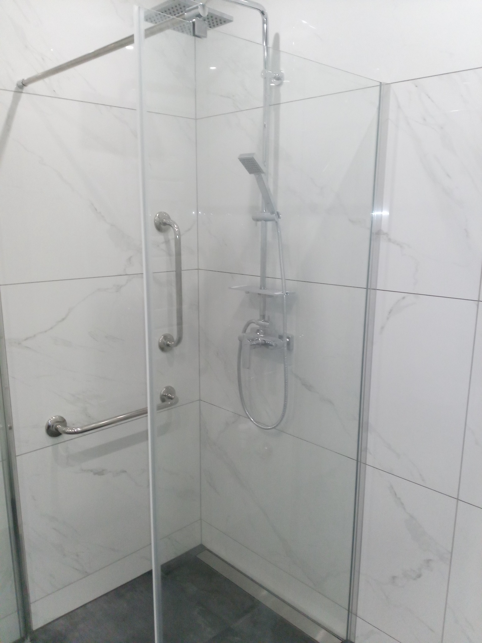Kabina prysznicowa z przeszklonymi drzwiami, chromowanymi uchwytami i deszczownicą, wykończona białymi płytkami imitującymi marmur, z szarym brodzikiem.