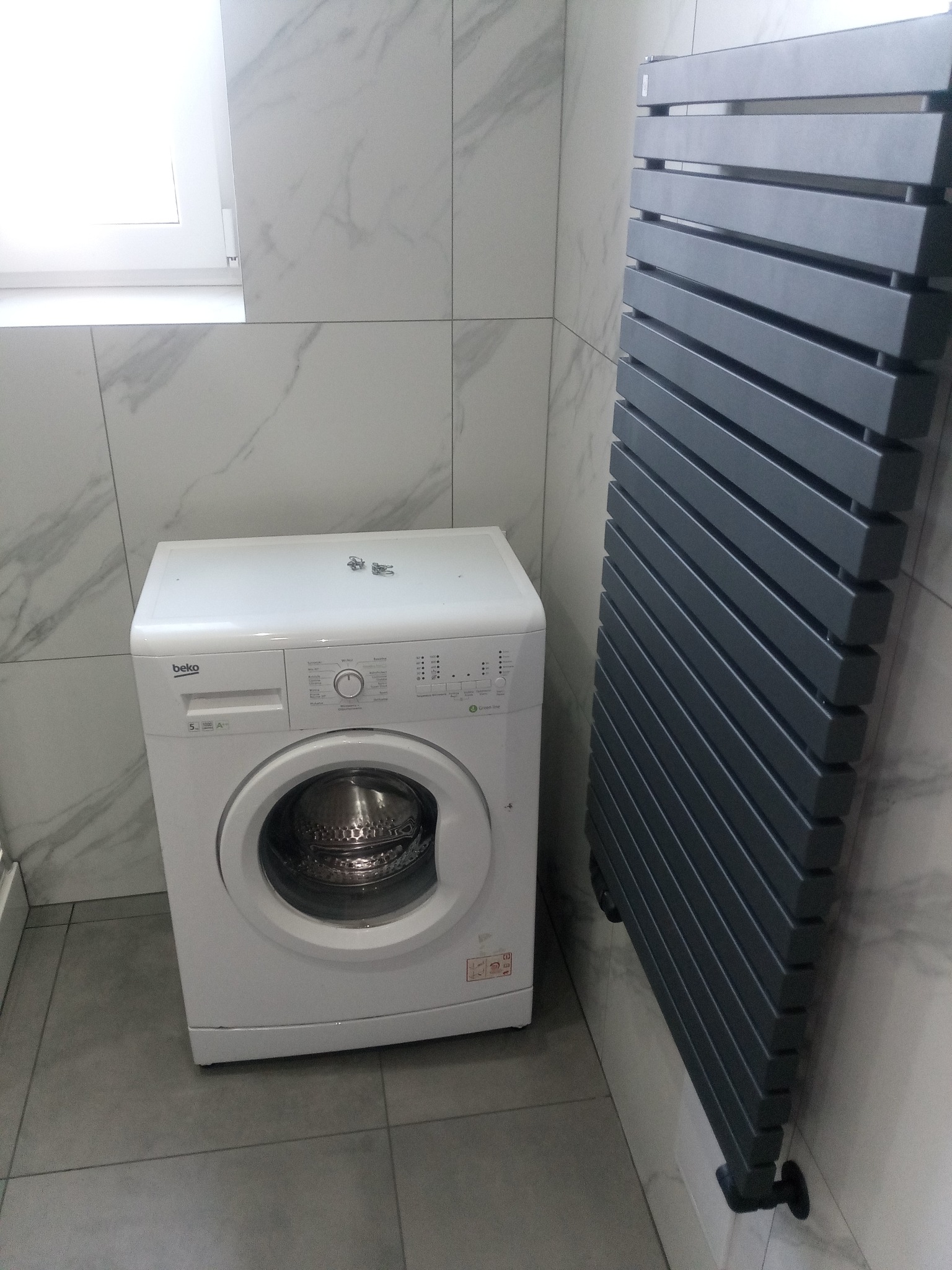 Nowoczesna łazienka z białą pralką Beko i grafitowym grzejnikiem drabinkowym na tle marmurowych płytek.