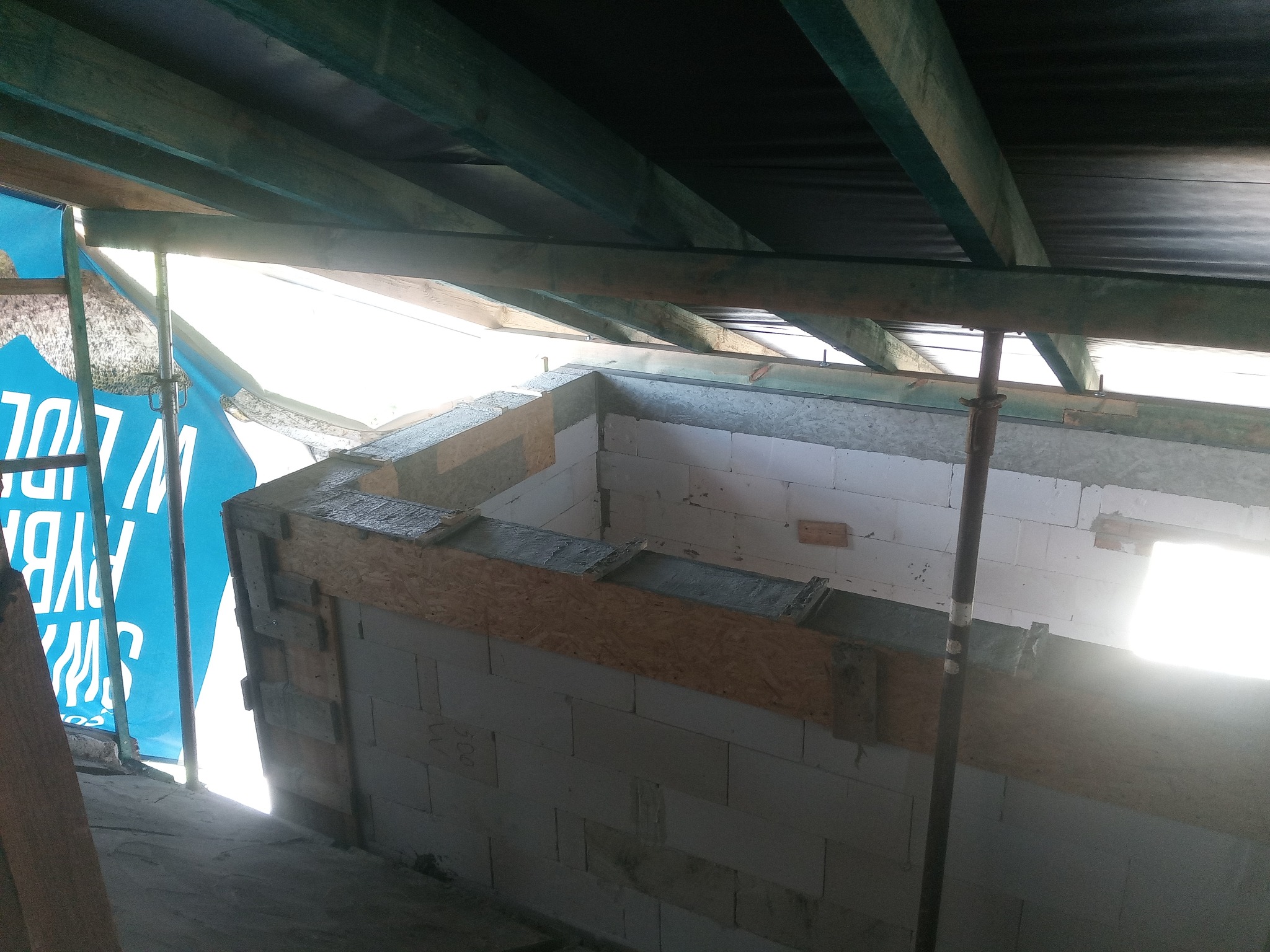 Wznoszenie ścian z bloczków betonowych z widocznym szalunkiem i zbrojeniem, fragment konstrukcji budynku w trakcie budowy, z widocznymi elementami drewnianymi więźby dachowej.