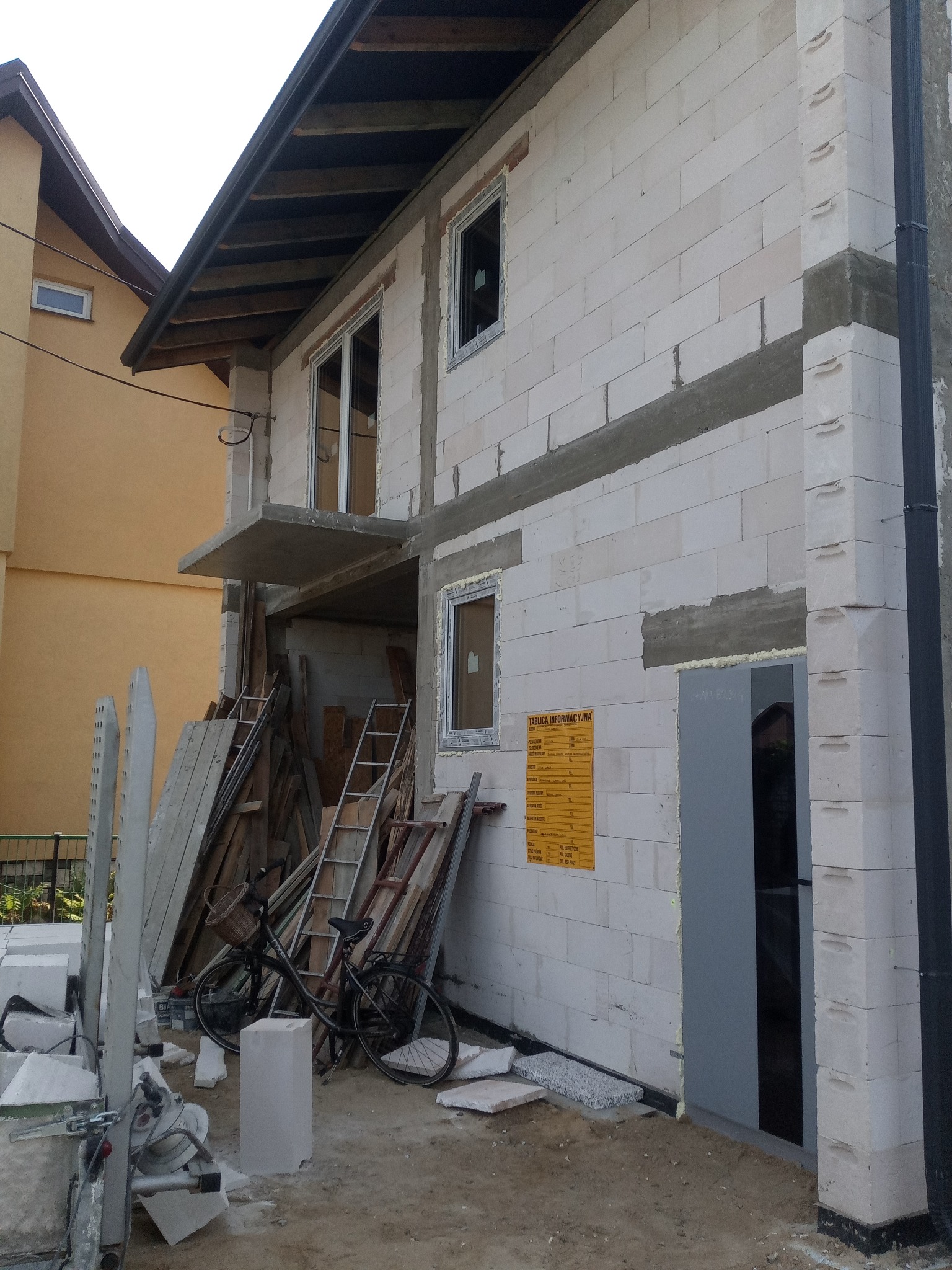 Surowy stan domu z białych bloczków, widoczne rusztowania, drabiny i rower z koszykiem na tle budowy.