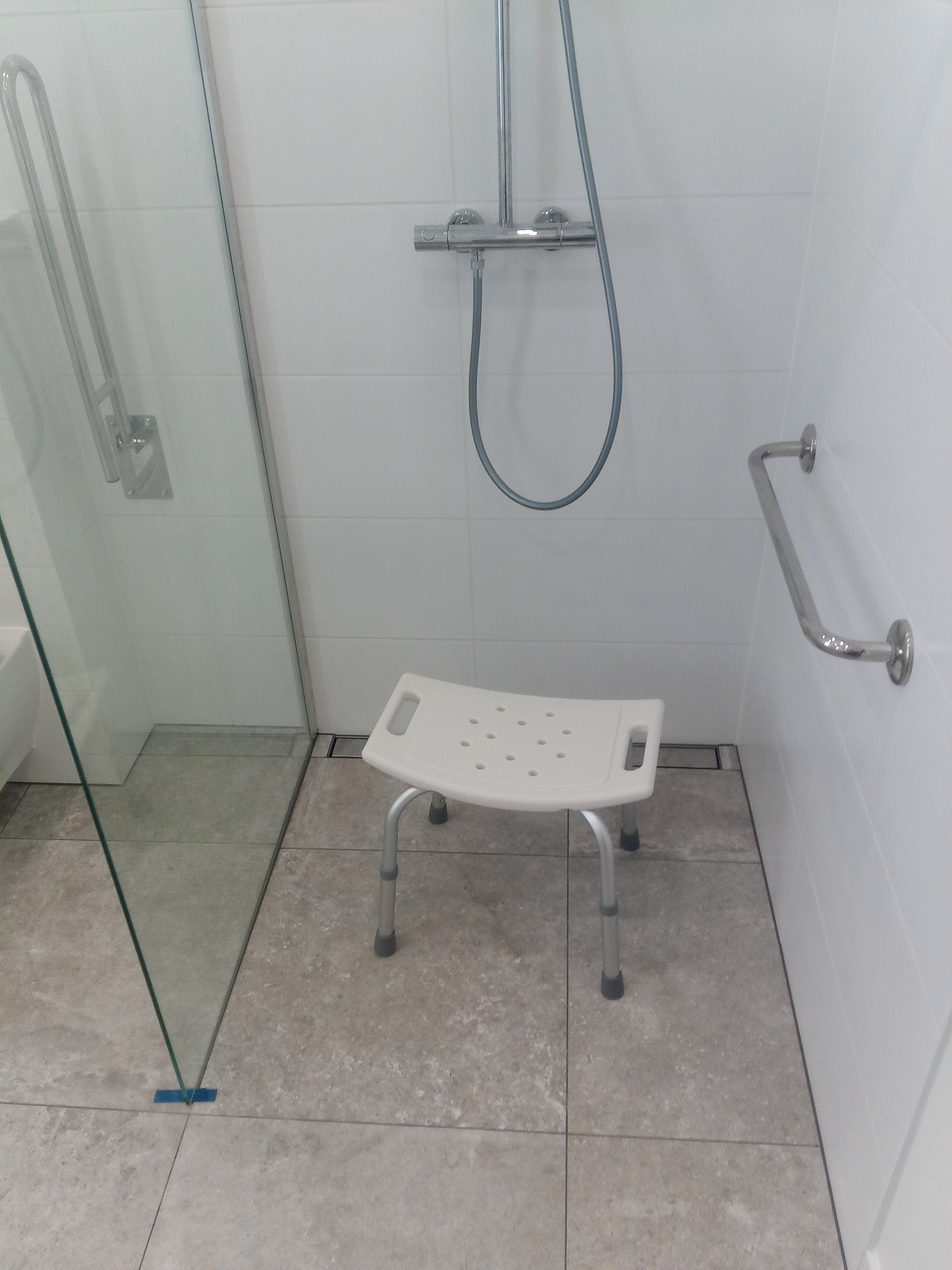 Biała łazienka z kabiną prysznicową, szarą podłogą, krzesłem prysznicowym z otworami i uchwytami dla osób z niepełnosprawnościami.