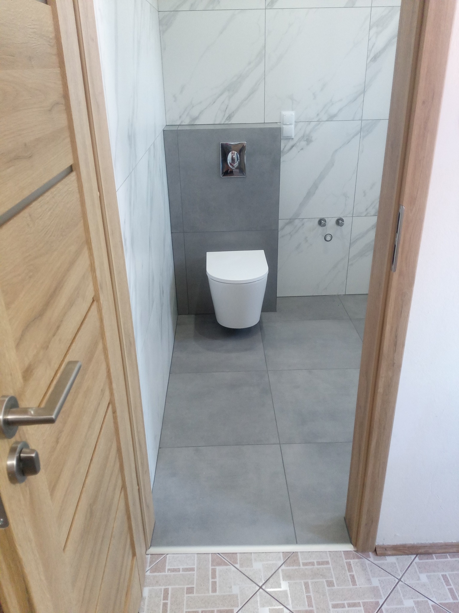 Nowoczesna łazienka z wiszącą miską WC, szarymi płytkami podłogowymi i marmurowymi płytkami ściennymi widziana zza częściowo otwartych drewnianych drzwi.