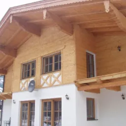 Dwupiętrowy dom z drewna z białym parterem, balkonem z drewnianą balustradą i zdobionym okapem, widoczne góry w tle.