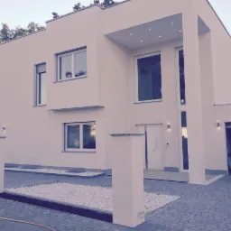 Nowoczesny, dwukondygnacyjny dom z płaskim dachem, białymi ścianami, dużymi oknami i elementami dekoracyjnymi z kamienia na podjeździe wyłożonym kostką brukową.