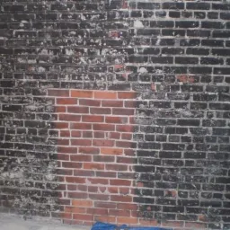 Częściowo oczyszczona ceglana ściana, fragmenty pomalowane na czarno, widoczne ślady wcześniejszych zabrudzeń i resztek farby, niejednolity kolor cegieł.