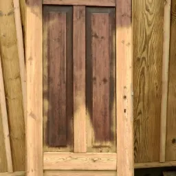 Częściowo odnowione drewniane drzwi z widocznymi śladami starej farby i surowego drewna, stojące na płycie OSB przed drewnianą ścianą.