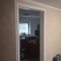 Wnętrze w trakcie prac, widok przez odnowione białe obramowanie drzwi, z widoczną ścianą z płytek po lewej i pomalowaną na beżowo ścianą po prawej stronie, w tle umeblowany pokój z oknem.