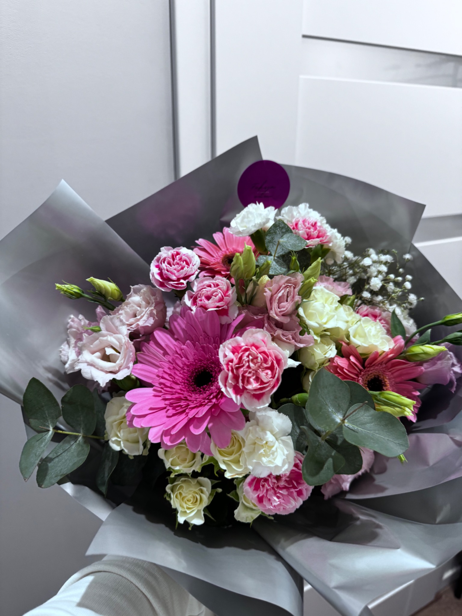 Bukiet mieszanych kwiatów w odcieniach różu i bieli, zapakowany w szary papier, z widoczną okrągłą fioletową naklejką.